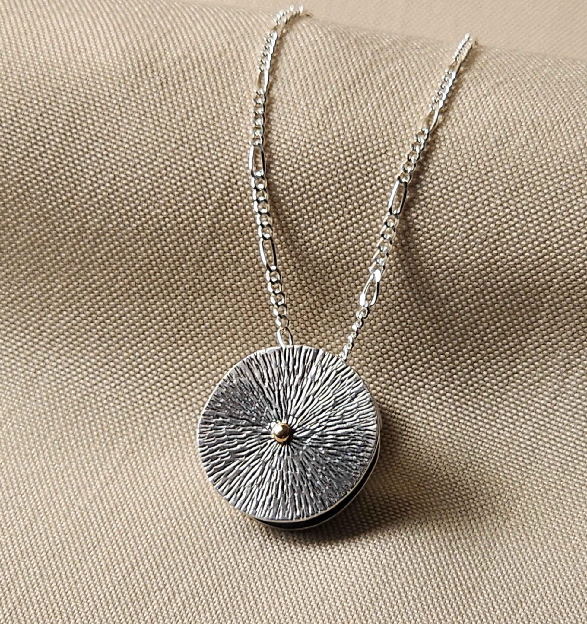 Silver necklace with a textured round pendant with 14k gold dot in center on a beige fabric background