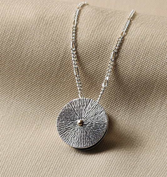 Silver necklace with a textured round pendant with 14k gold dot in center on a beige fabric background